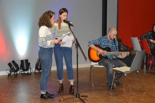 Two Young women are Standing in front of a microphone singing. Next to them a Young man is sitting on a chair playing the guitare. 